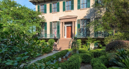 Image Of A Tan Colored House With Dark Green Shutters And A Lush Garden In The Foreground.